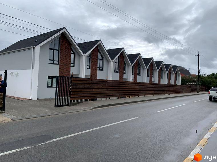 Таунхауси Scandi House з двосхилими дахами та цегляними вставками, розташовані вздовж дерев'яного паркану. Зовнішній вигляд новобудови та прибудинкової території.