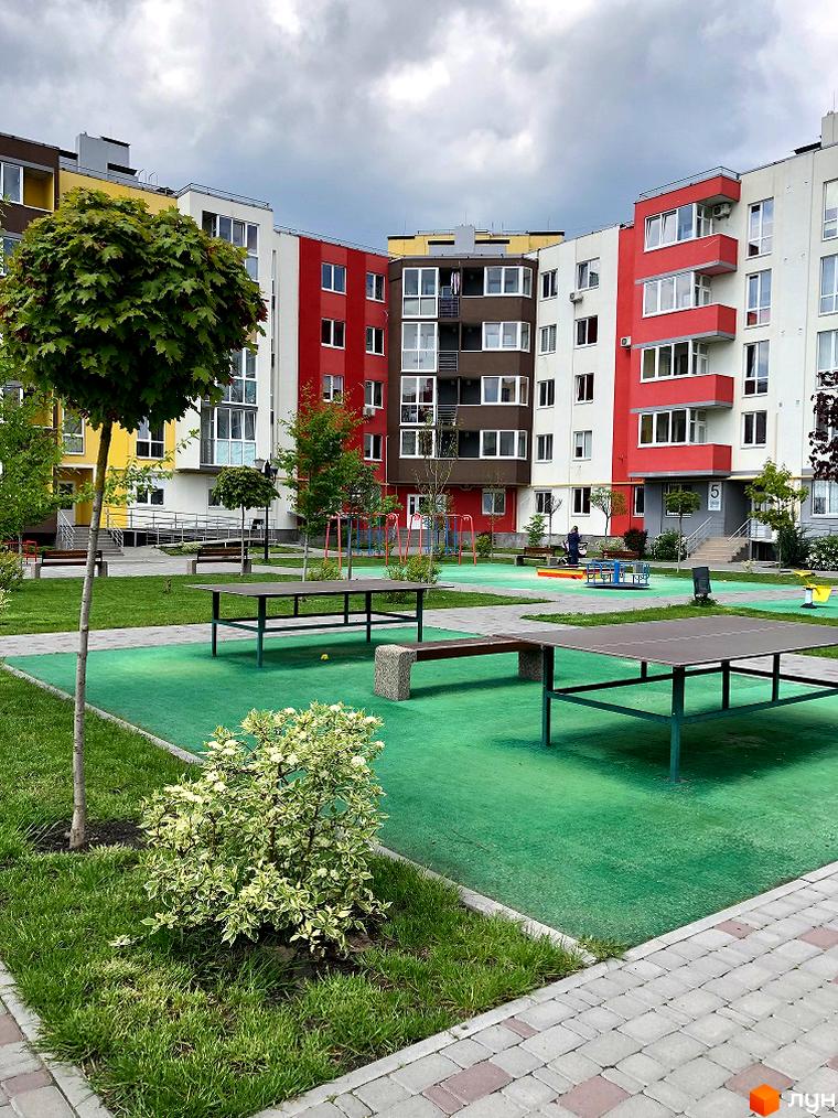 Житловий комплекс Нова Буча: фасади будинків з червоними та білими елементами, дитячий майданчик з гойдалками та каруселлю, столи для настільного тенісу.