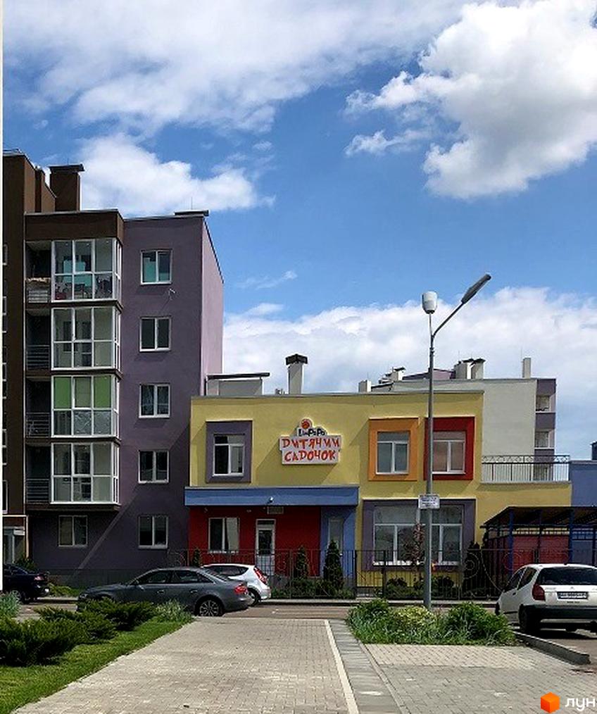 Житловий комплекс Нова Буча. Фасад будинку з вікнами та балконами. Навколо будинку зелена зона та припарковані автомобілі.