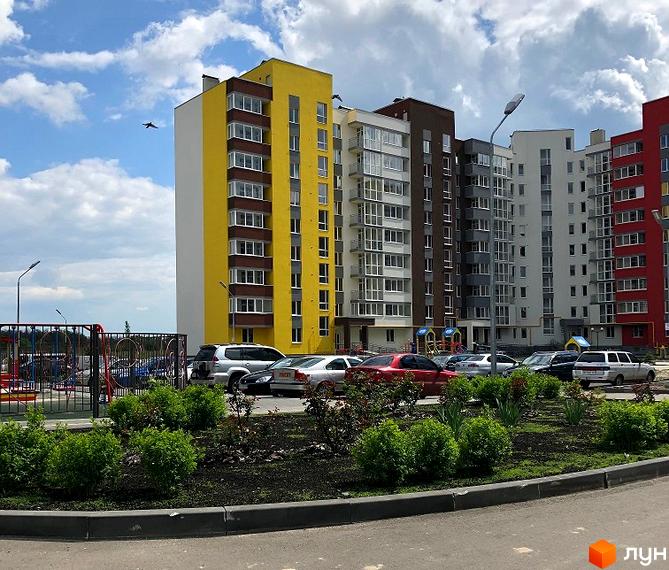 Житловий комплекс Нова Буча: яскраві фасади будинків та зелена прибудинкова територія з дитячим майданчиком. Парковка з автомобілями поруч із зеленими насадженнями.