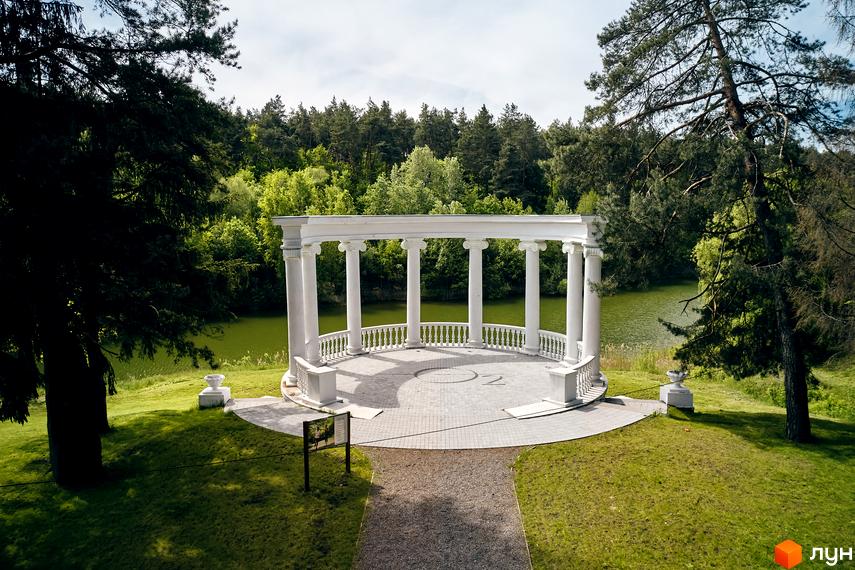 Житловий комплекс O2 Residence: біла ротонда з колонами на березі озера, оточена деревами. Доглянута територія з газоном та доріжками.