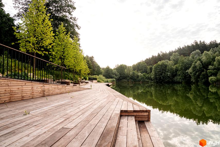 O2 Residence: дерев'яний настил біля озера з зеленою рослинністю та відображенням хмар у воді. Зона відпочинку на березі водойми.