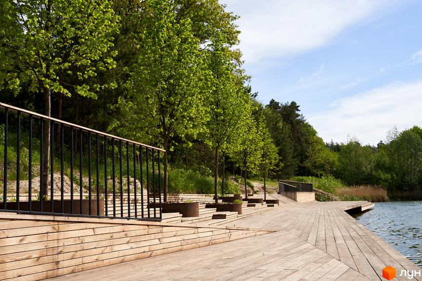Житловий комплекс O2 Residence: дерев'яний настил біля озера з зеленими деревами та огорожею. Територія комплексу з ландшафтним дизайном.