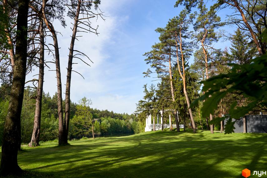 Житловий комплекс O2 Residence: біла альтанка з колонами на зеленій галявині серед сосен. Паркова зона з доглянутим газоном та тінями від дерев.