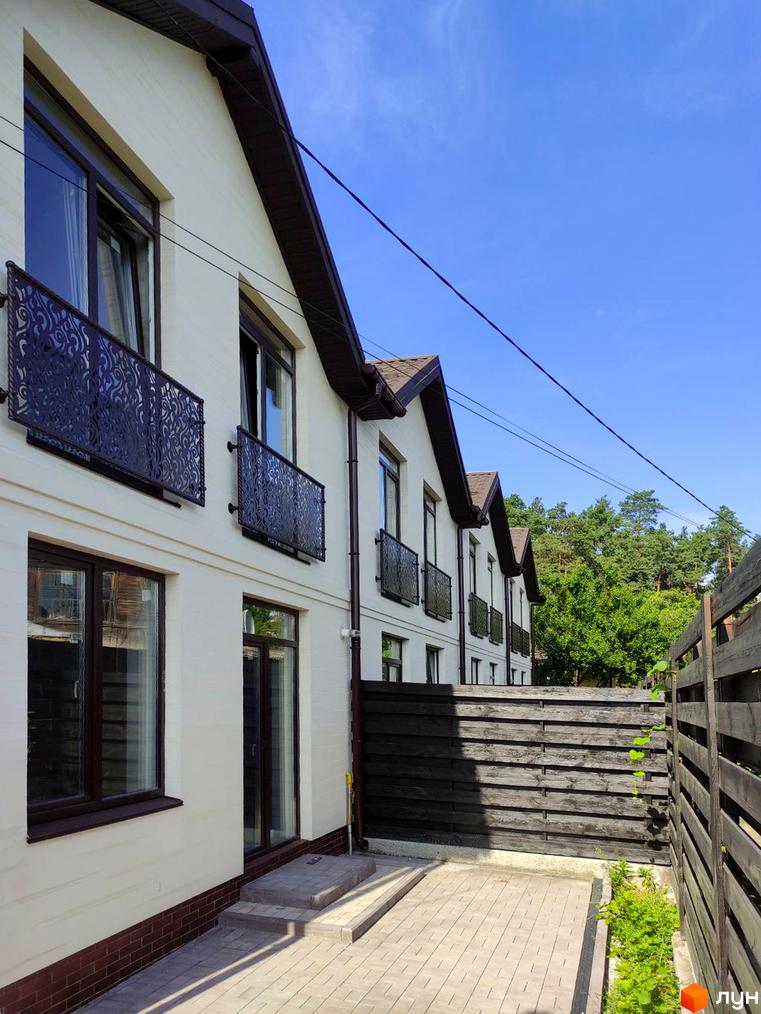 Ряд білих таунхаусів Travertino з коричневими дахами та вікнами, прикрашеними кованими балконами. Перед будинками вимощений двір та дерев'яний паркан, на задньому плані ліс.