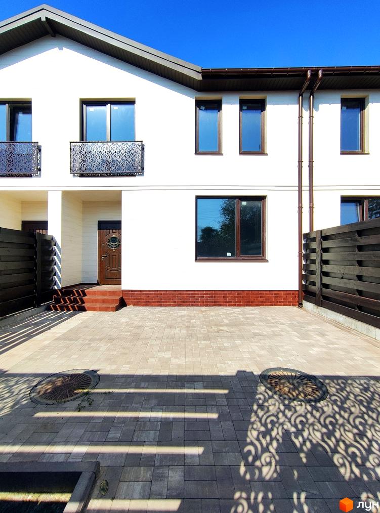 Білий таунхаус Travertino з коричневими вікнами та декоративними балконами. Прибудинкова територія викладена бруківкою, є дерев'яні огорожі.