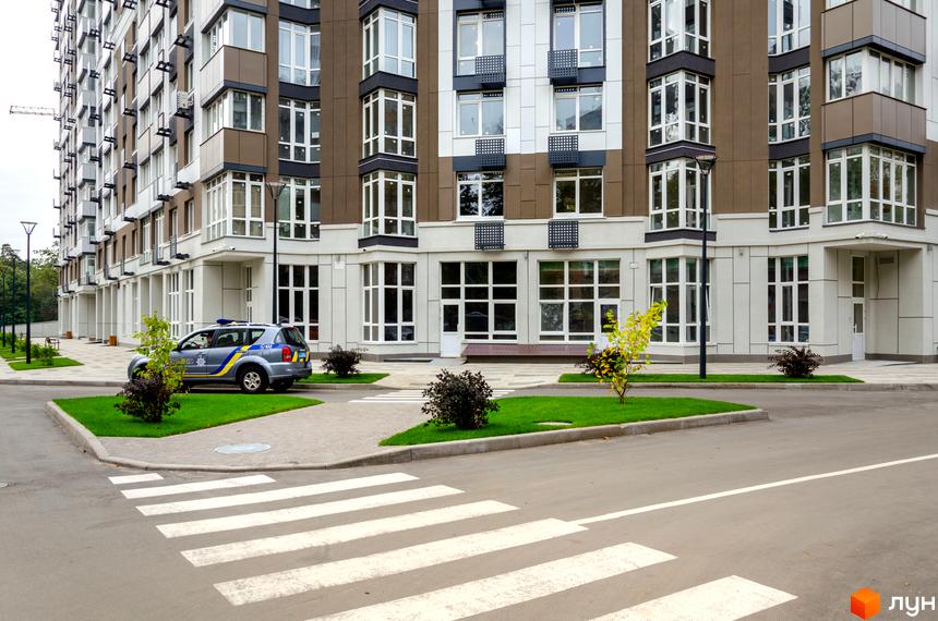 Зовнішній вигляд новобудови ЖК Святобор з поліцейським автомобілем та пішохідним переходом. Прибудинкова територія з газоном та деревами.