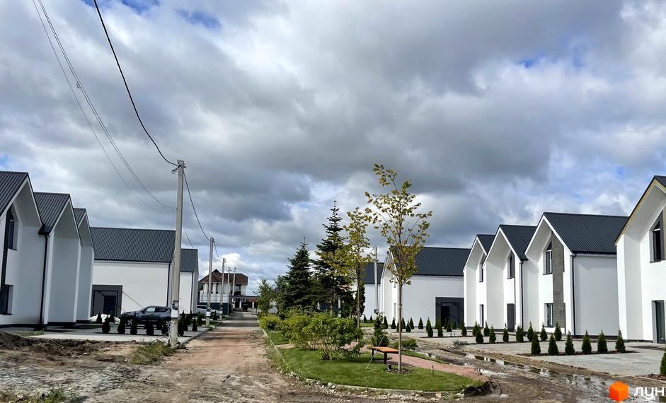 Котеджне містечко Щаслива Вулиця: білі двоповерхові будинки з темними дахами. Прибудинкова територія має зелені насадження та ґрунтову дорогу.