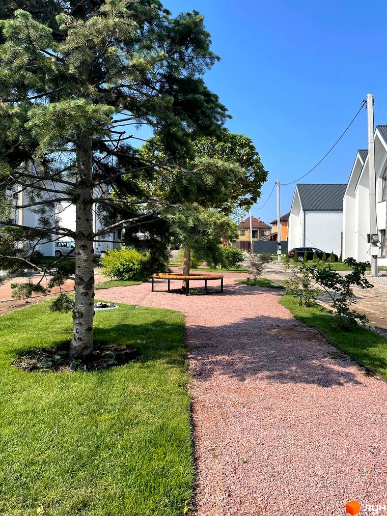 Котеджне містечко Щаслива Вулиця: білі будинки з темними дахами, оточені зеленими газонами та доріжками з гравію, з деревами та лавкою.