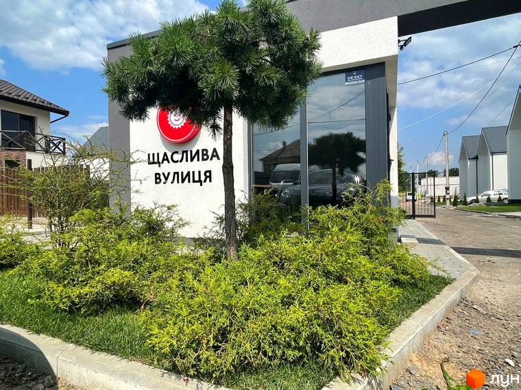 Котеджне містечко Щаслива Вулиця: фасади будинків та зелена прибудинкова територія з деревами та кущами.