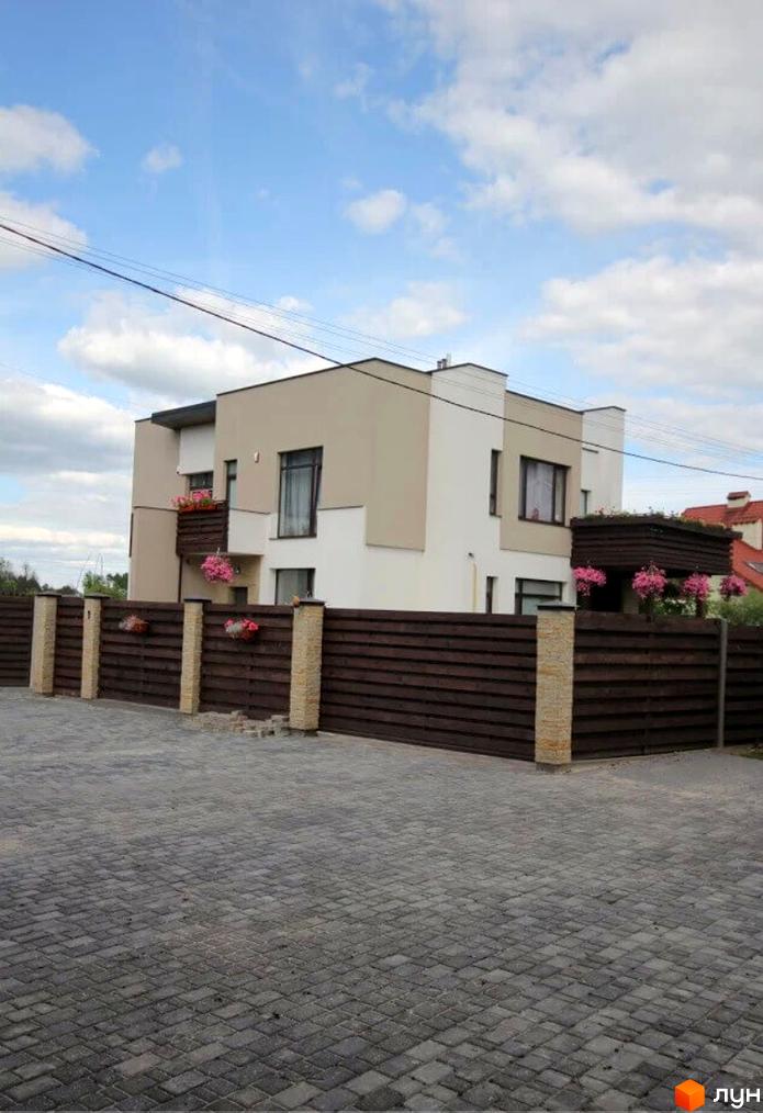 Котеджне містечко КМ Life: двоповерховий будинок з бежевим та білим фасадом, огороджений дерев'яним парканом з кам'яними стовпами. Навколо будинку облаштована бруківка.