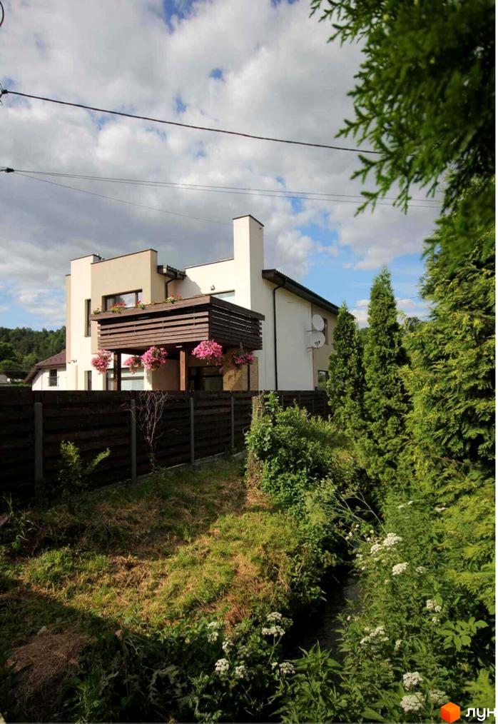 Котеджне містечко КМ Life: двоповерховий будинок з дерев'яним балконом та квітами, огороджена територія з деревами та зеленню.