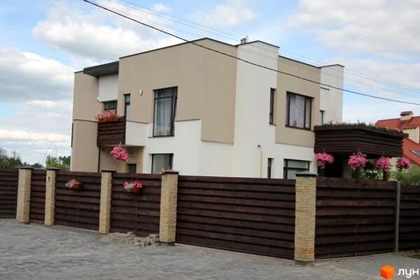 Зовнішній вигляд будинку КМ Life з коричневим парканом та квітами. Прибудинкова територія з бруківки та паркану.