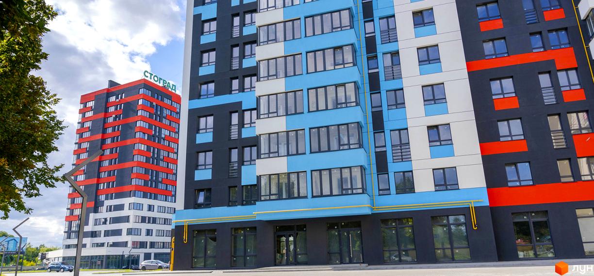 Фасад житлового комплексу Bridge Town з яскравими червоними та блакитними елементами, що доповнюється зеленою зоною та дорогою з автомобілями.