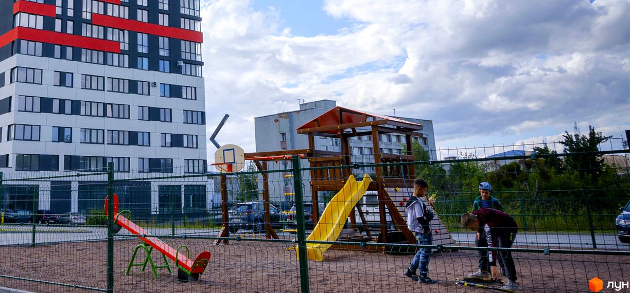 Житловий комплекс Bridge Town: багатоповерховий будинок з червоними та сірими фасадами, дитячий майданчик з гіркою та гойдалкою.