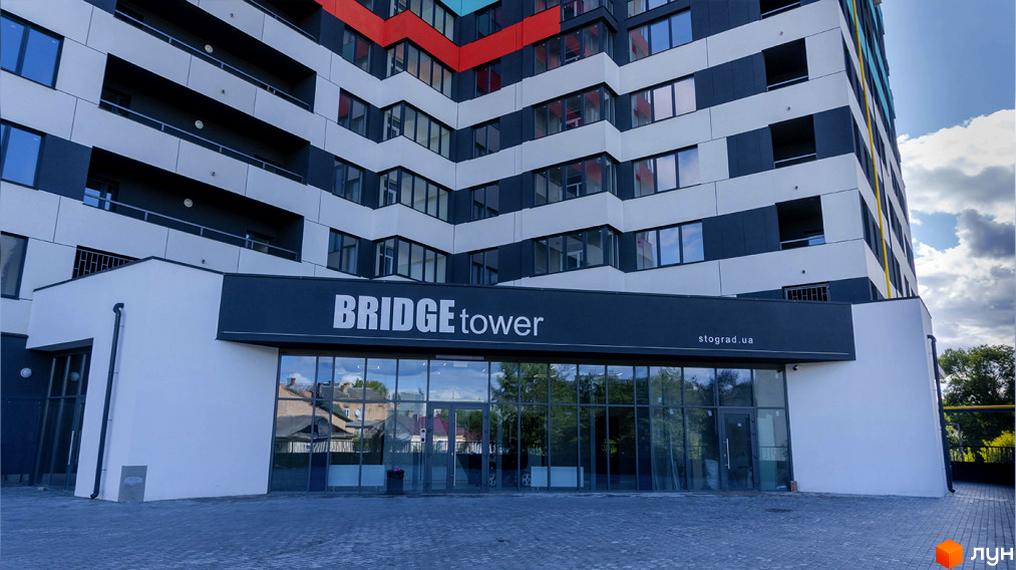 Новобудова Bridge Tower із скляним фасадом та балконами. Перед будинком облаштований вхід із великими вікнами та бруківкою.