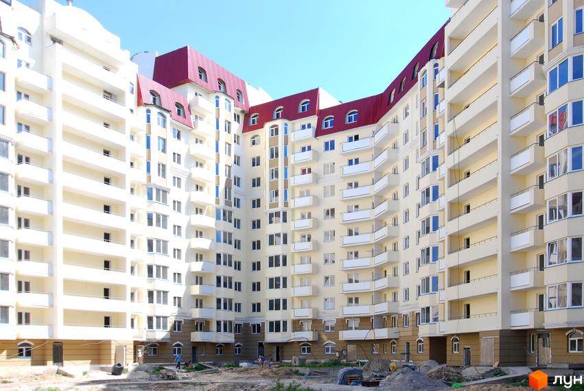 Багатоповерховий житловий комплекс Західний зі світлим фасадом, червоним дахом та численними балконами. Прибудинкова територія знаходиться на етапі облаштування.