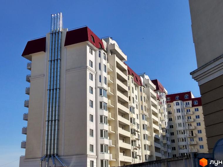 Багатоповерховий будинок Житлового комплексу Західний з світлим фасадом, червоним дахом та балконами. На фасаді видно вертикальні металеві труби.