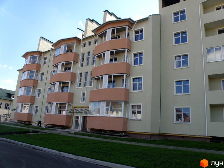 Житловий комплекс Петропавлівський з фасадом світлого кольору, вікнами та округлими балконами. Перед будинком зелений газон та дорога.