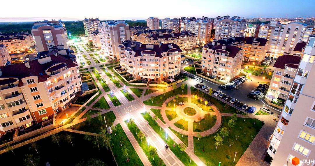 Житловий комплекс Петрівський квартал. Багатоповерхові будинки з балконами та вікнами, освітлені ввечері. Навколо будівель — зелені зони з доріжками та освітленням.