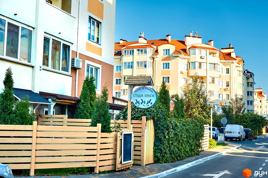 Житловий комплекс Петрівський квартал з фасадами світло-бежевого кольору та червоними дахами. Прибудинкова територія огороджена дерев'яним парканом та озеленена туями.