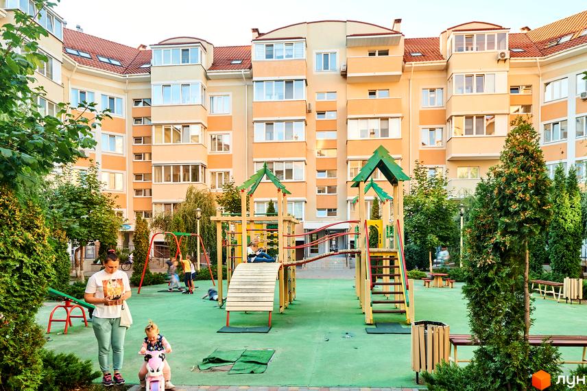 Житловий комплекс Петрівський квартал. Будинки світло-бежевого кольору з червоними дахами. На прибудинковій території дитячий майданчик з гірками та гойдалками.