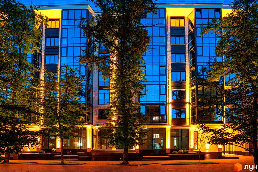 Житловий комплекс Royal Hall: фасади з панорамним склінням та підсвічуванням, оточені деревами. Територія перед будинком вимощена плиткою.