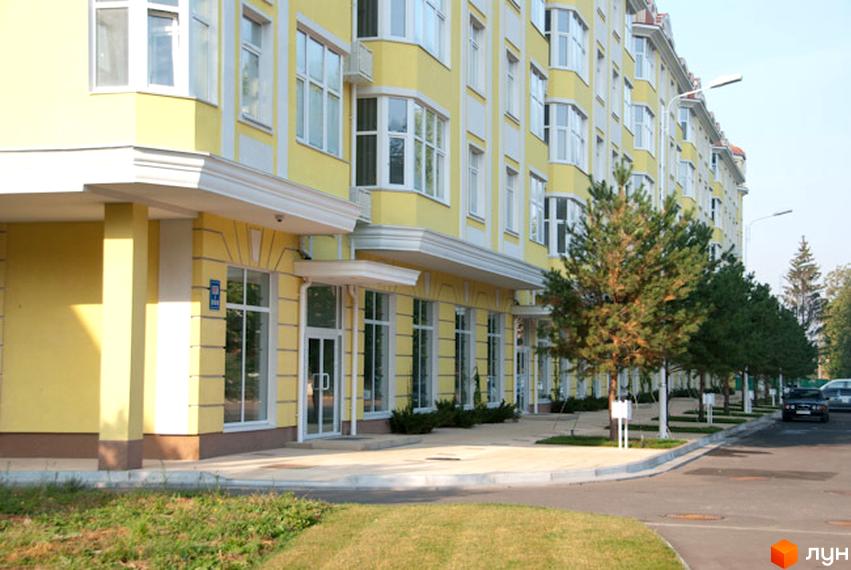 Зовнішній вигляд будинку ЖК Щасливе з жовтою фасадною обробкою та великими вікнами. Прибудинкова територія озеленена деревами та газонами.