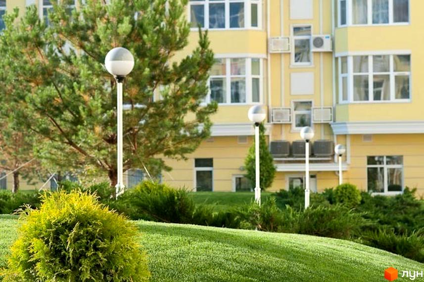 Житловий комплекс Щасливе з жовтою фасадною частиною та прибудинковою територією з зеленими насадженнями та ліхтарями.