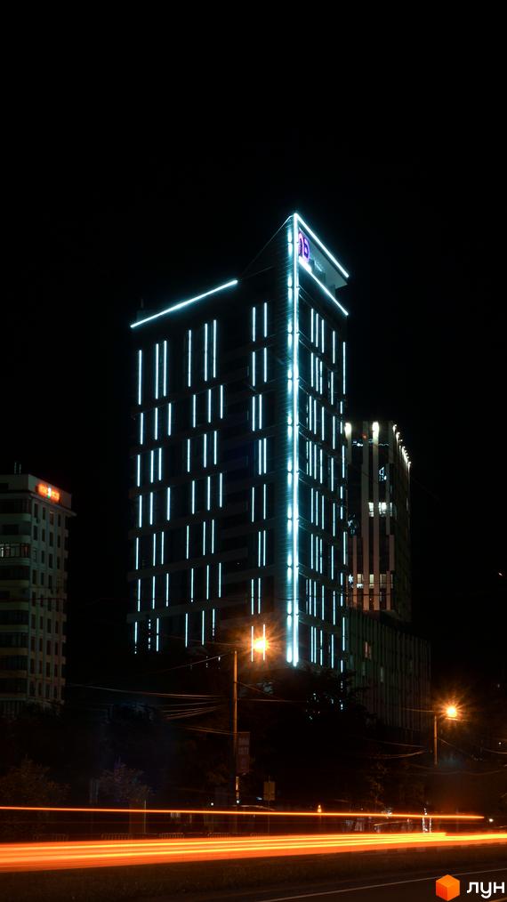 Житловий комплекс Щасливий Platinum у нічний час з неоновим освітленням фасаду. Навколо будівлі видно вуличне освітлення та смуги від фар автомобілів на дорозі.