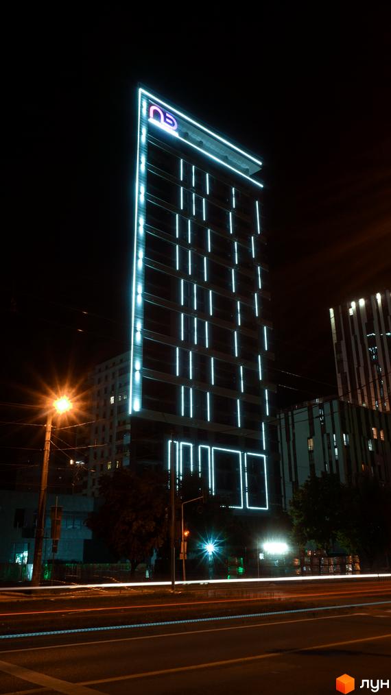 Новобудова ЖК Щасливий Platinum вночі з неоновим підсвічуванням фасаду. Зовнішній вигляд будівлі з вертикальними світловими елементами та логотипом проєкту.