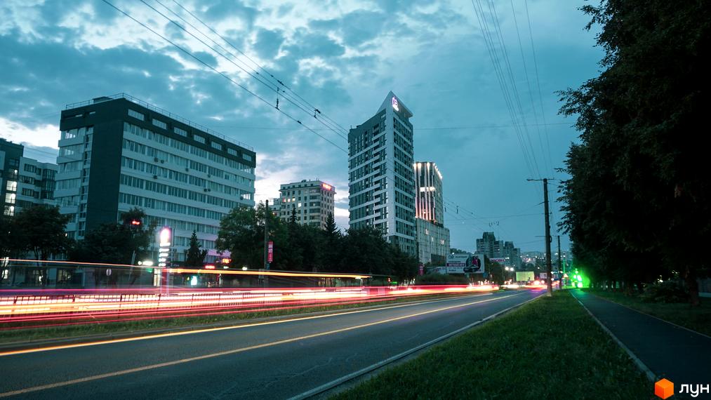 Новобудова ЖК Щасливий Platinum з фасадом з панорамних вікон та сучасним дизайном. Вечірній вид на міський проспект з освітленими будівлями та зеленою зоною.