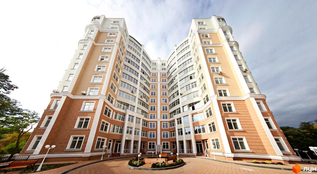 Висотна будівля житлового комплексу з великою кількістю вікон. На прибудинковій території розташовані лавки та клумби.