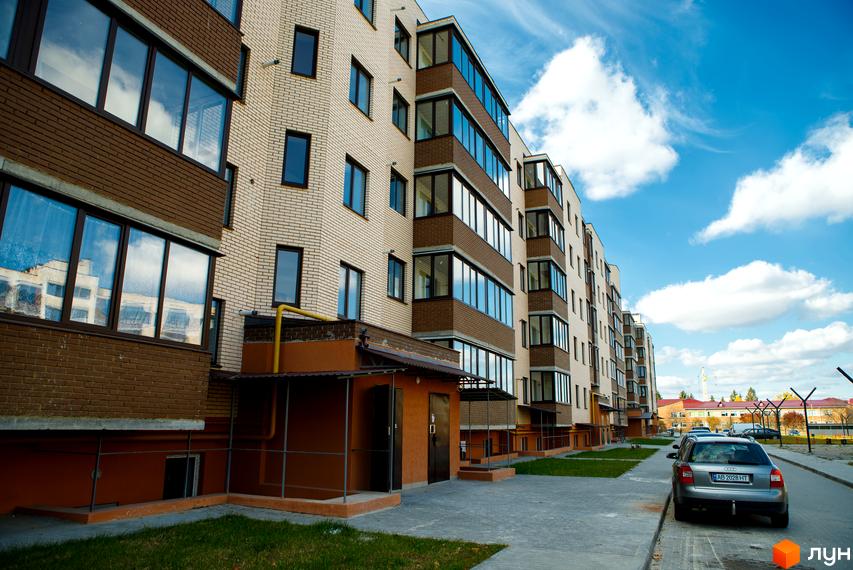 Зовнішній вигляд новобудови ЖК Софія з цегляним фасадом та скляними балконами. Прибудинкова територія з тротуаром та припаркованими автомобілями.