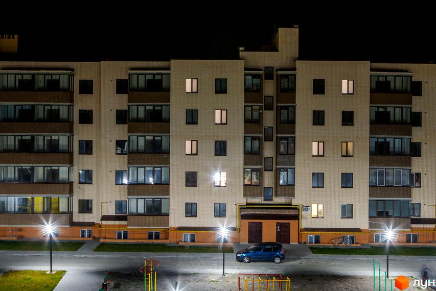 Новобудова ЖК Софія вночі. Багатоповерховий будинок з вікнами та балконами. На прибудинковій території є освітлення, дорога та дитячий майданчик.