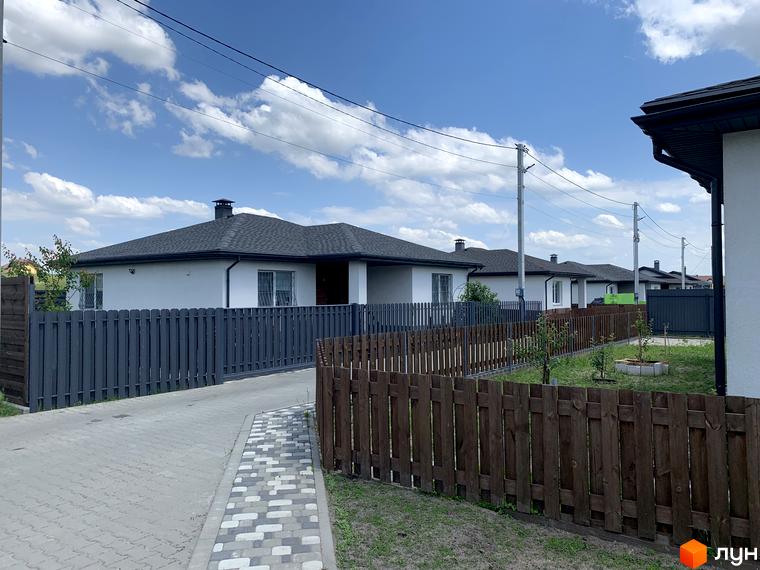 Котеджне містечко Тарасівка: білі одноповерхові будинки з темними дахами та парканами. На території облаштована бруківка та зелені насадження.