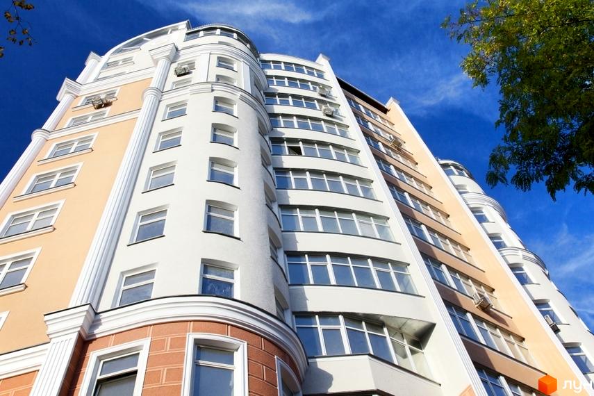 Висотна будівля житлового комплексу з великою кількістю вікон на фоні блакитного неба. Фасад будинку виконаний у світлих тонах з декоративними елементами.