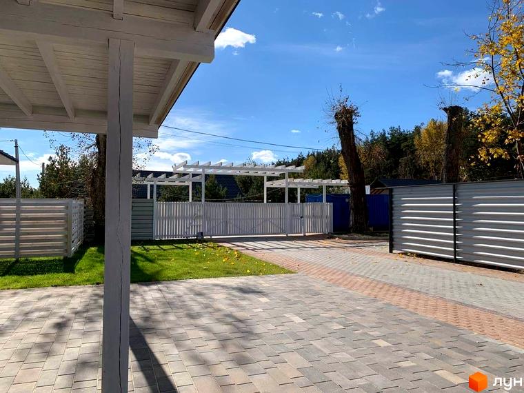 Котеджне містечко КМ Wood Side Village з білими парканами та вимощеною доріжкою. Навколо зелена трава та дерева.