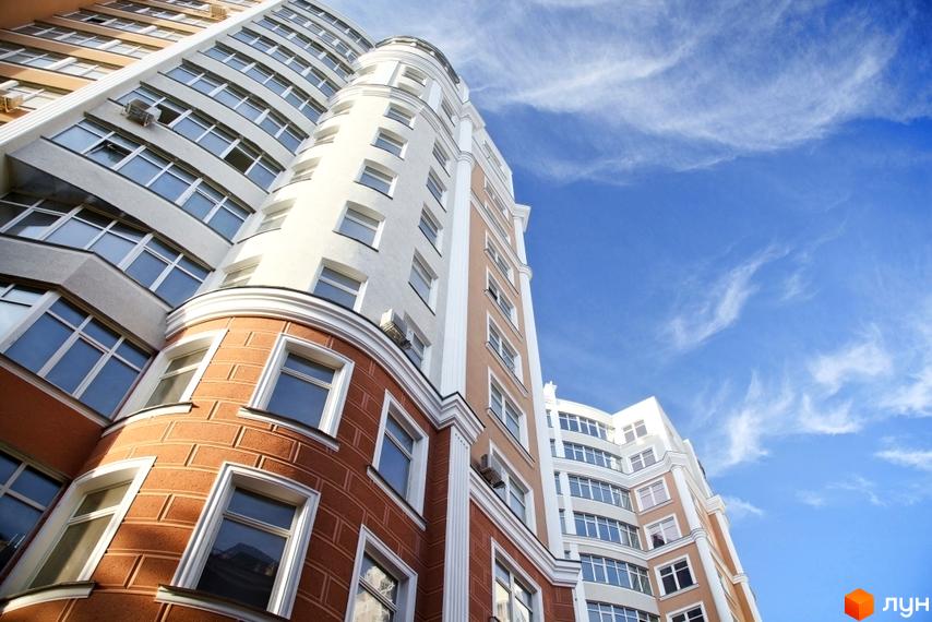 Висотна будівля житлового комплексу з великою кількістю вікон на фоні блакитного неба. Фасад будівлі виконаний у світлих тонах з елементами коричневого кольору.