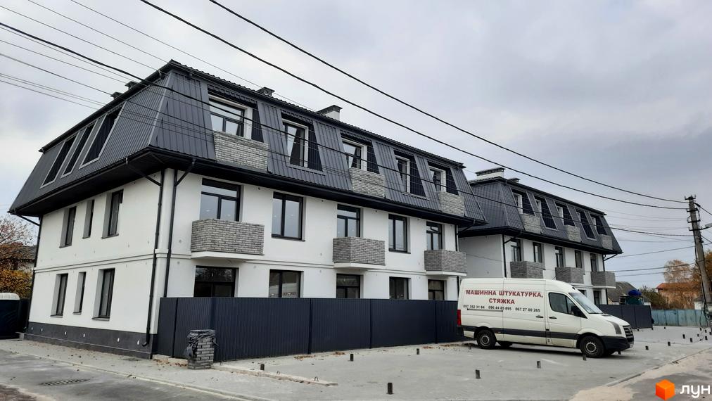 Білі багатоповерхові будинки з темно-сірими мансардними дахами та балконами. Перед ними розташована вимощена територія з парканом.