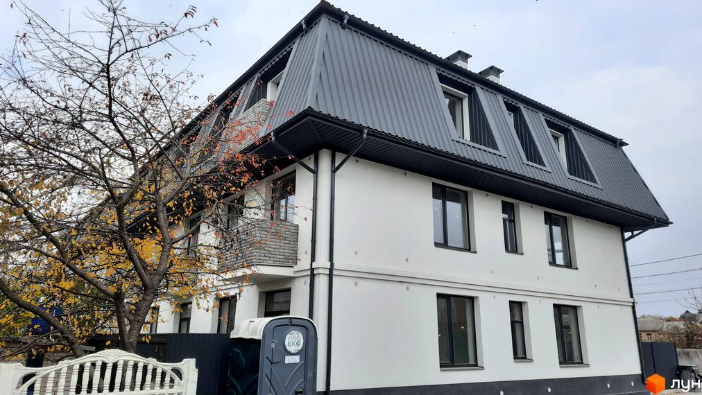 Зовнішній вигляд триповерхового будинку з сірим дахом та білими стінами на вулиці Вчительська, 16. Поруч розташоване дерево з жовтим листям та портативна вбиральня.