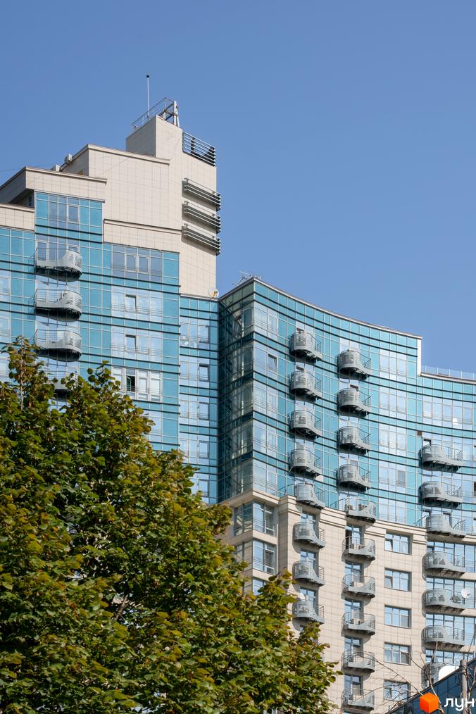 Житловий комплекс Парус має вигнутий скляний фасад з численними балконами та світлими вставками. На передньому плані видно зелене дерево.