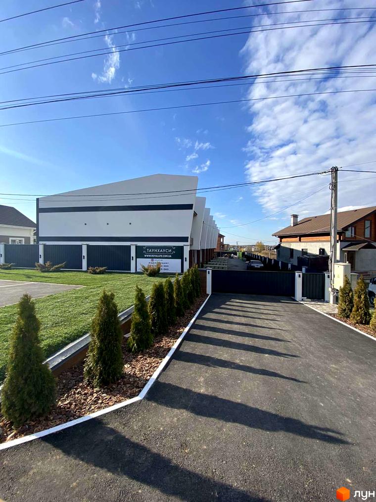 Таунхауси Royal life – це комплекс з білими фасадами та чорними елементами, розташований на доглянутій території з акуратними зеленими насадженнями та асфальтованою дорогою.