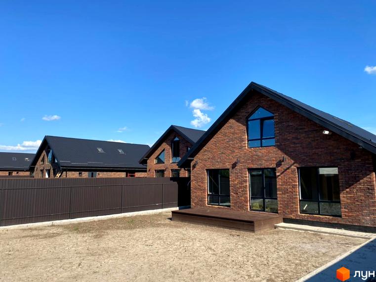 Котеджне містечко Одноповерховий Київ - Гореничі. Будинки з цегляними фасадами, темними дахами та великими вікнами. Перед будинками ділянка землі та дерев'яна тераса.