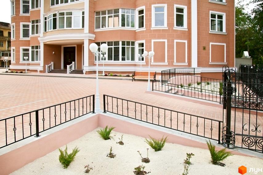 Зовнішній вигляд будинку житлового комплексу з пісочницею та кованими огорожами. На території комплексу висаджені рослини.