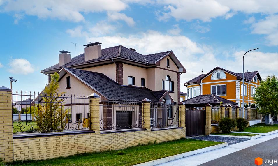 Котеджне містечко КМ Home Resort: двоповерхові будинки світло-бежевого та жовтого кольорів з темними дахами, огороджені цегляним парканом з кованими елементами. Прибудинкова територія з газоном та деревами.