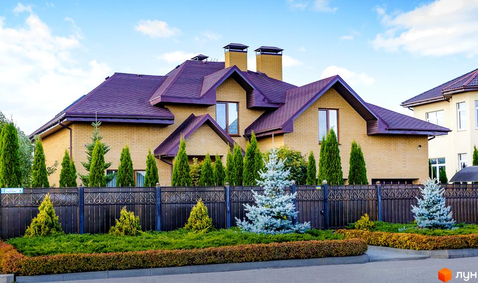 Котеджне містечко КМ Home Resort. Будинок з жовтої цегли з фіолетовим дахом, оточений туями та декоративними ялинками. Огороджена територія з кованою огорожею та доглянутим ландшафтом.