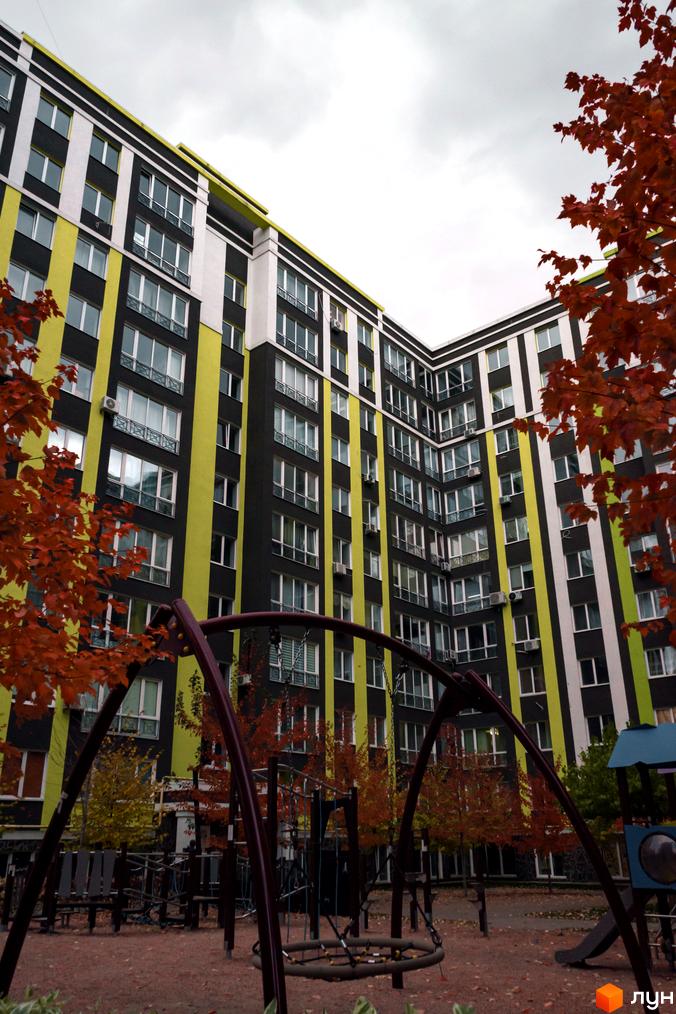 Житловий комплекс Фортуна-2 з фасадом у чорно-жовтих тонах. На передньому плані дитячий майданчик з гойдалками та осінніми деревами.