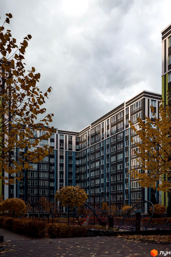 Висотна будівля ЖК Фортуна-2 з великою кількістю вікон. На території комплексу видно дерева з жовтим листям та дитячий майданчик.