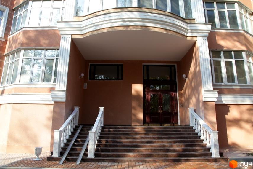 Вхідна група житлового комплексу з мармуровими перилами та сходами. Фасад будівлі виконаний у коричневих тонах з великими вікнами.